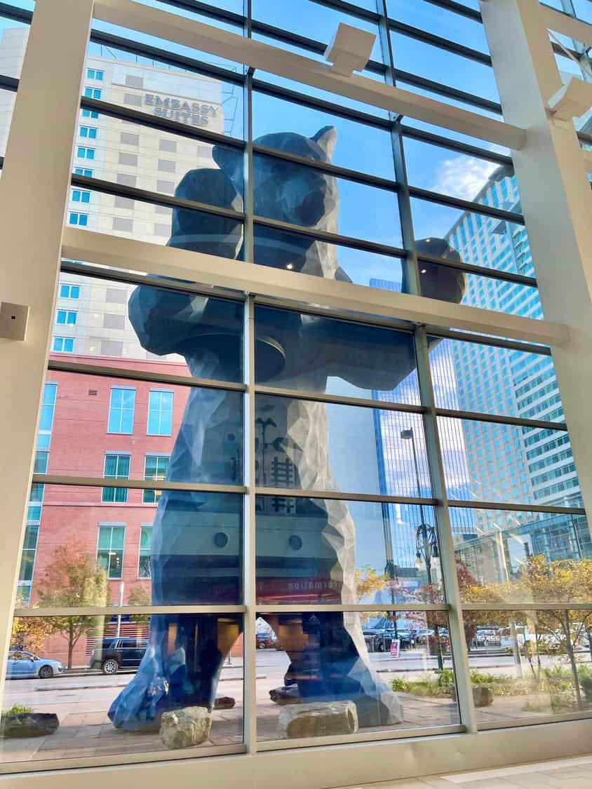 A giant faceted blue bear sculpture presses its paws against a multi-pane glass atrium, visible from inside a downtown street lined with high-rise buildings; sign reads "EMBASSY SUITES".