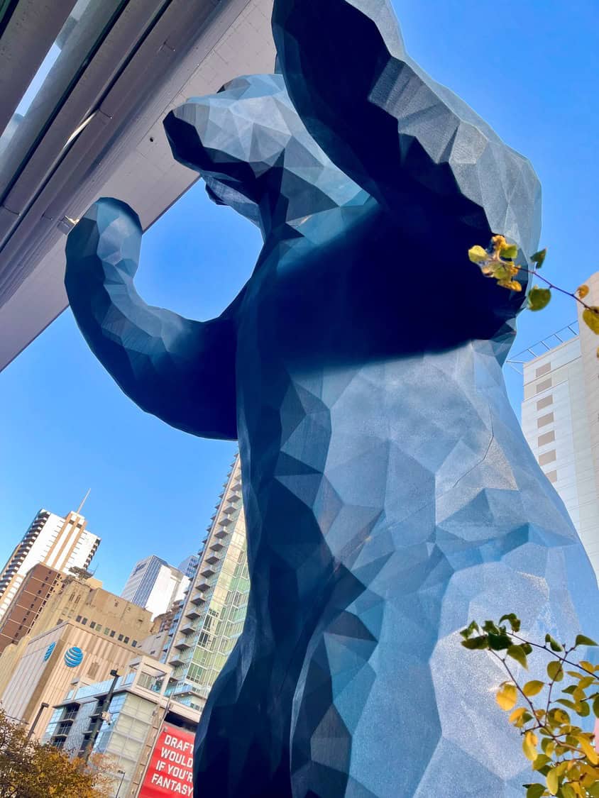 Large faceted blue bear sculpture rearing with paws raised, towering among downtown skyscrapers beneath a clear blue sky. 
Text visible: "AT&T" (logo) and red billboard reading "DRAFT WOULD IF YOUR FANTASY".