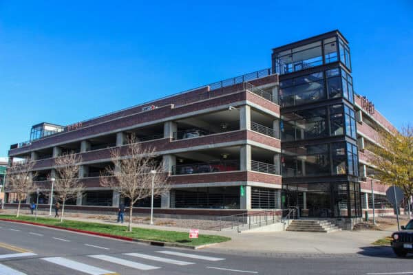 Multi-level brick-and-concrete parking garage holds parked cars; glass-enclosed stair tower at the corner provides access; pedestrians walk along the sidewalk with leafless trees by crosswalk under clear blue sky.