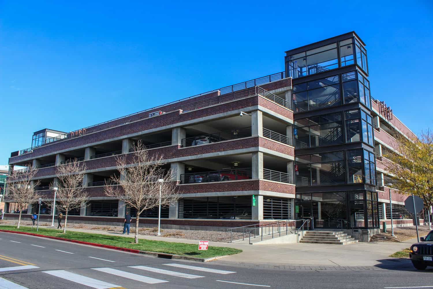 Multi-level brick-and-concrete parking garage holds parked cars; glass-enclosed stair tower at the corner provides access; pedestrians walk along the sidewalk with leafless trees by crosswalk under clear blue sky.