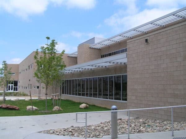 Beige brick building shows long glass windows beneath metal sunshades, set beside a concrete walkway with planted trees and decorative rocks on a sunny suburban campus.