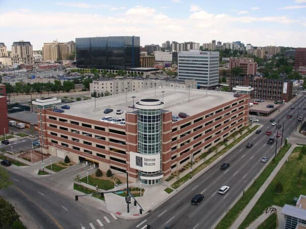A multi-story parking garage bearing a "DENVER HEALTH" sign, holding parked cars on rooftop and ramps, bordered by a multi-lane street with moving vehicles and surrounding commercial buildings.

Text found: "DENVER HEALTH"; "VP"