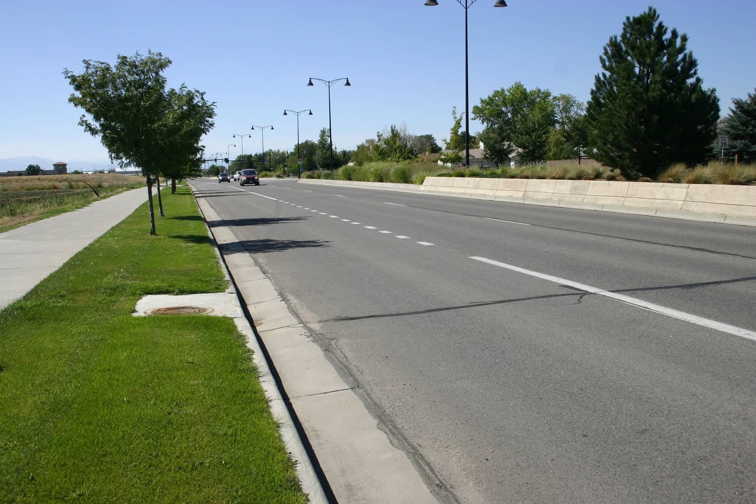 Wide asphalt road carries a few cars toward the distance; concrete median and lined streetlights separate lanes; grassy sidewalk with trees runs alongside; clear blue sky. Sign reads "35".