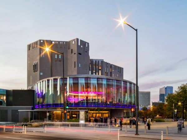 Round glass theater glows with purple interior lighting, hosting people at its entrance, set against a blocky concrete tower and city street at dusk with lit streetlamps.