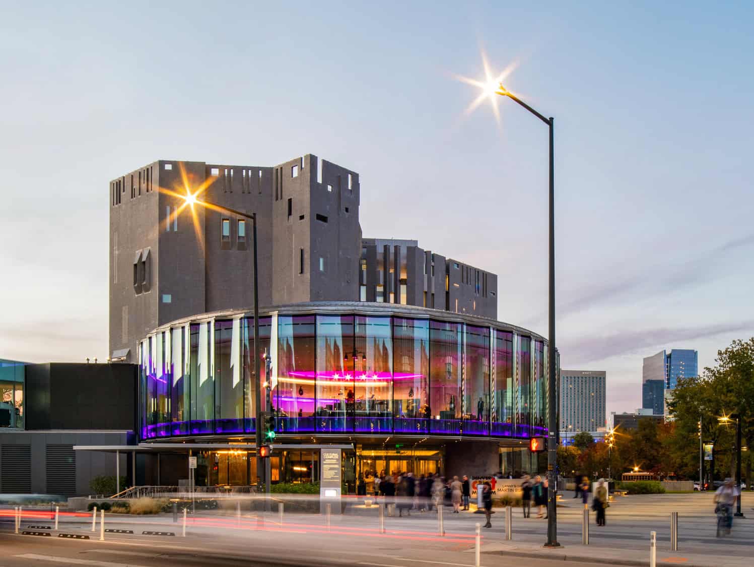 Round glass theater glows with purple interior lighting, hosting people at its entrance, set against a blocky concrete tower and city street at dusk with lit streetlamps.