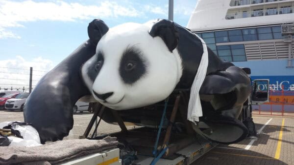 Large panda sculpture resting on a flatbed trailer, strapped for transport in a port parking lot beside a docked cruise ship and rows of parked cars.

Visible text: "RoyalC"  "ERNA"