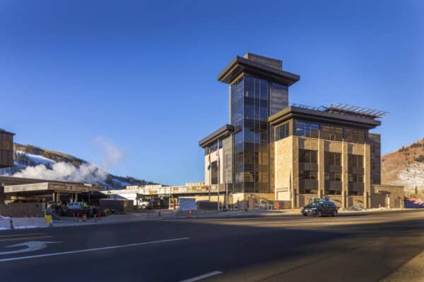 A multi-story glass-and-stone building stands beside a road; construction vehicles and workers operate at its base; snow-covered hills and clear blue sky frame the scene.