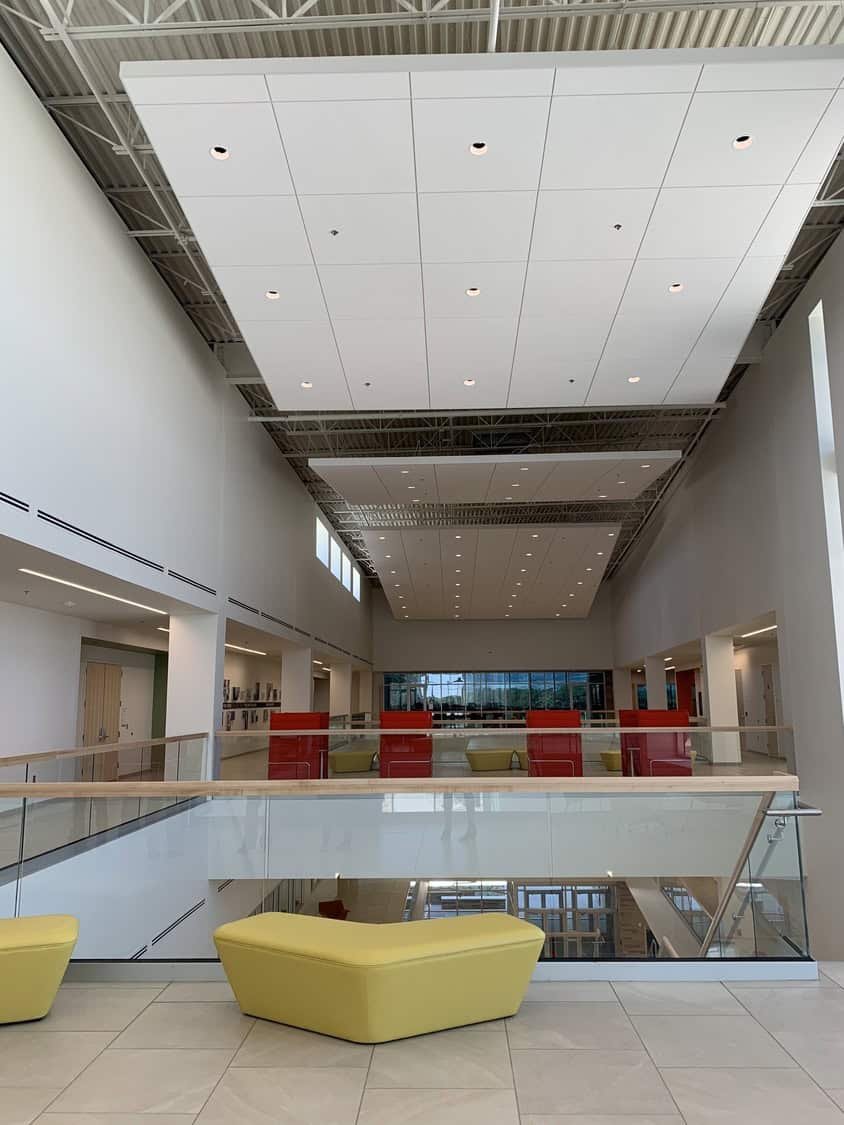 Yellow angular benches sit near a glass railing overlooking a lower level; red high-back chairs line the far walkway under large white suspended ceiling panels in a bright, minimalist atrium.