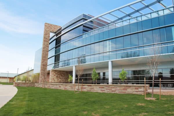 Curved glass-and-stone office building rises, its reflective windows mirroring the blue sky; young trees and a manicured lawn sit in front, with outdoor seating beneath an overhang.
