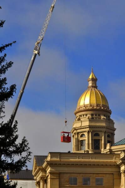 Crane hoisting a red work basket carrying two workers beside a gold-domed neoclassical building under a partly cloudy blue sky.