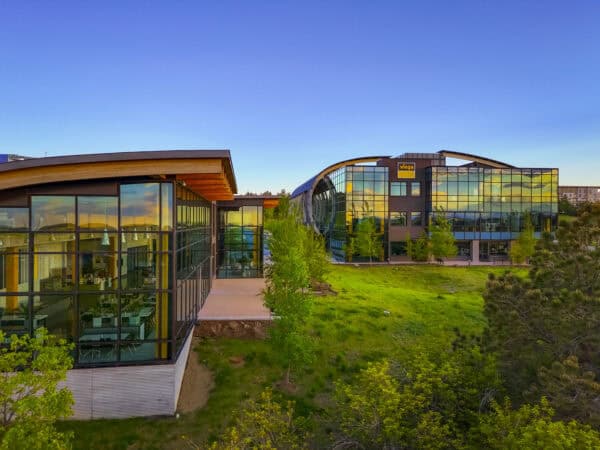 Two glass office buildings reflect sunset light; left building reveals interior dining area, right building displays viega sign. Grassy lawn and young trees sit in the foreground beneath a clear blue sky.