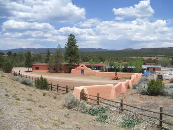 A low pink adobe wall and single-story buildings enclose a compound beside a dirt road and wooden fence, surrounded by scrub, pines and distant mountain ridges under a cloudy sky.