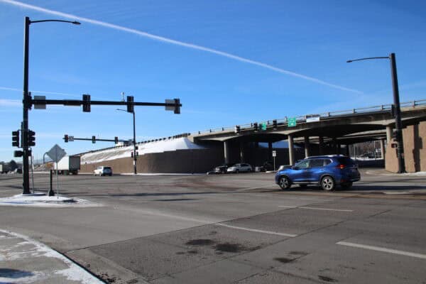 Blue SUV crosses a wide intersection, passing under a concrete highway overpass with traffic lights overhead, clear blue sky and light patches of snow on curbs and embankments.