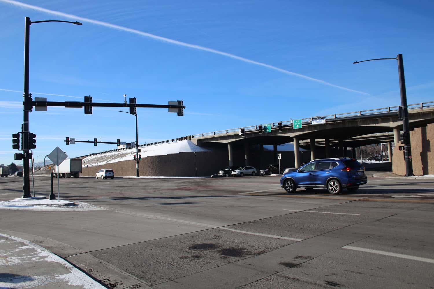 Blue SUV crosses a wide intersection, passing under a concrete highway overpass with traffic lights overhead, clear blue sky and light patches of snow on curbs and embankments.