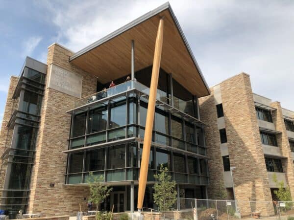 Stone-and-glass academic building — features a tall slanted wooden support and people on an upper balcony — framed by a partly cloudy sky and temporary construction fencing.

Text: "MICHAEL SMITH NATURAL RESOURCES BUILDING"