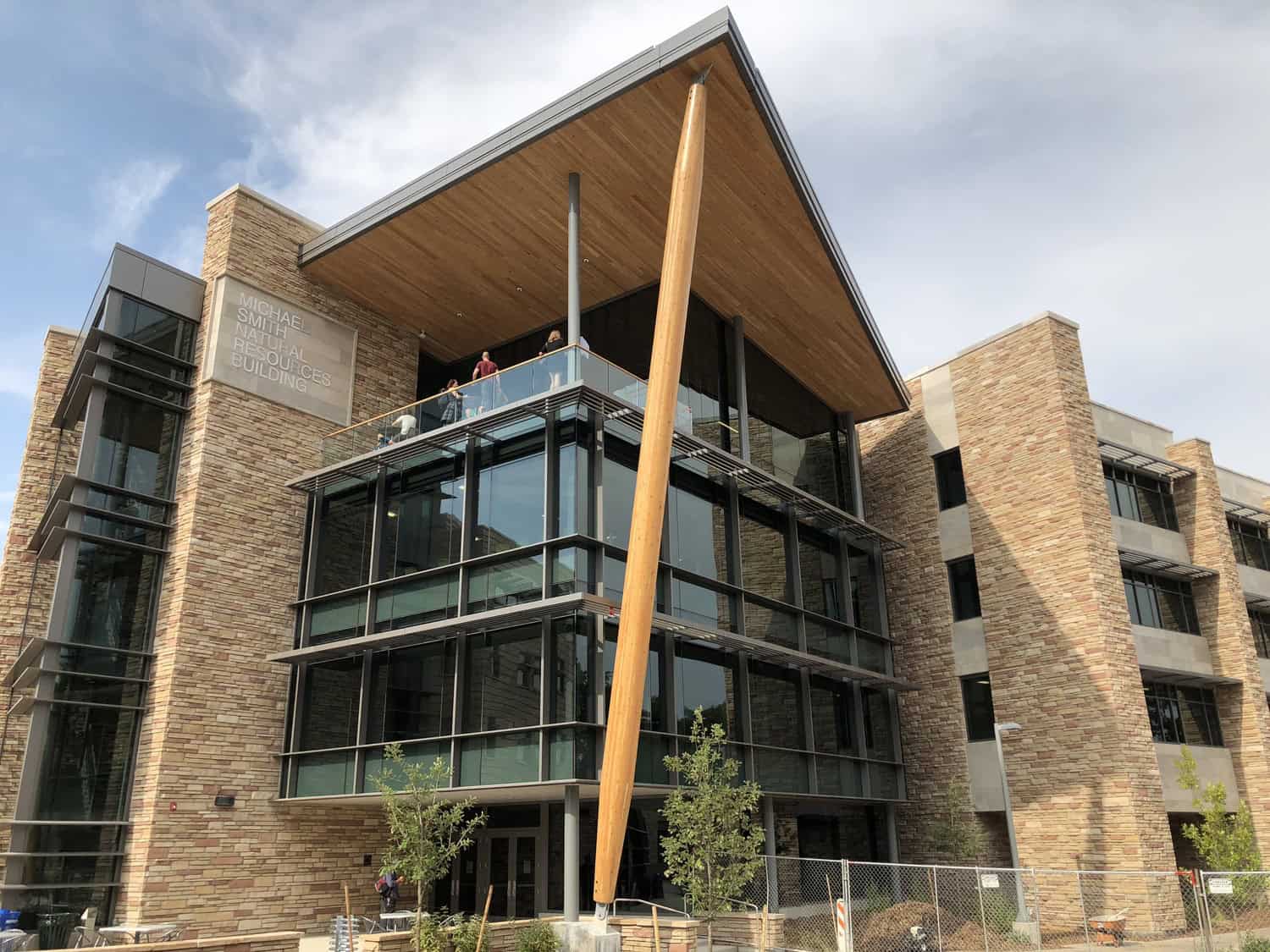 Stone-and-glass academic building — features a tall slanted wooden support and people on an upper balcony — framed by a partly cloudy sky and temporary construction fencing.

Text: "MICHAEL SMITH NATURAL RESOURCES BUILDING"