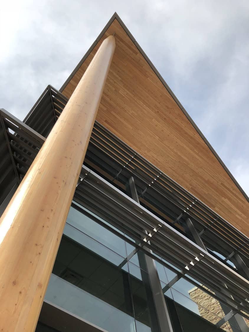 A tall round wooden column rises, supporting a wide angled wooden roof; metal sunshades and glass curtain-wall below reflect sky and adjacent stonework, photographed from a low-angle exterior view.