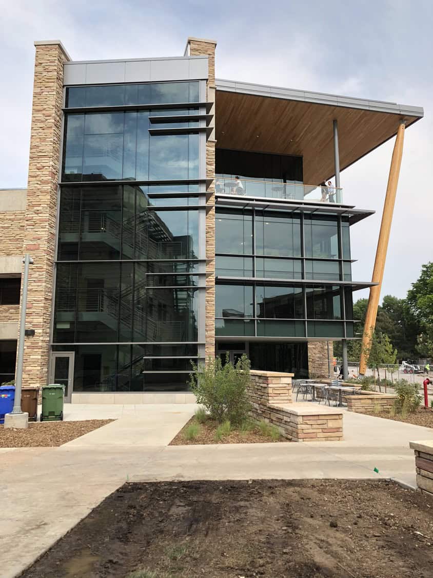 Modern glass-and-stone building with expansive curtain-wall windows; several people stand on a second-floor balcony; concrete plaza with planted beds and an angled wooden support beam in front.