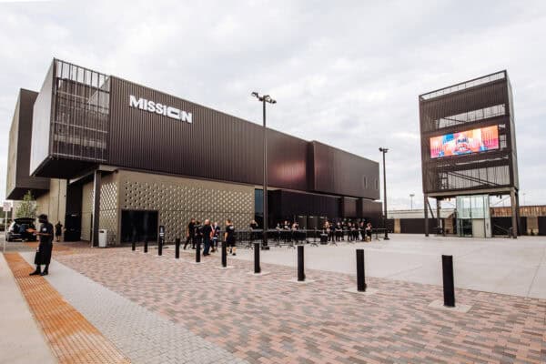 Modern black building labeled "MISSION" with staff managing a queue at its entrance on a wide paved plaza; tall tower displays colorful sign reading "MISSION LAUNCH" under a cloudy sky.