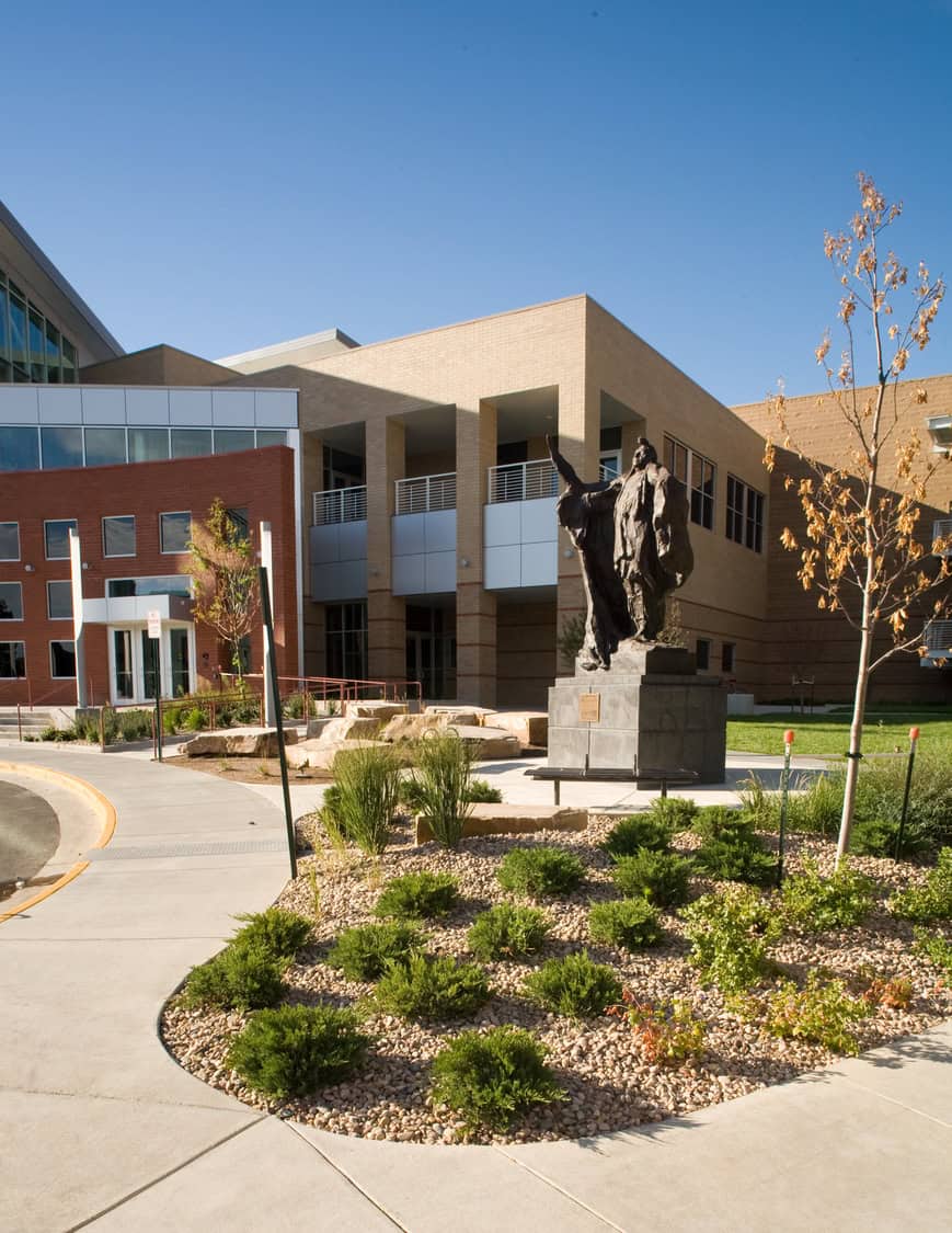 Bronze statue gestures upward atop a square stone pedestal; it stands in a gravel-and-shrub landscaped courtyard; a modern brick-and-glass academic building and clear blue sky surround it.