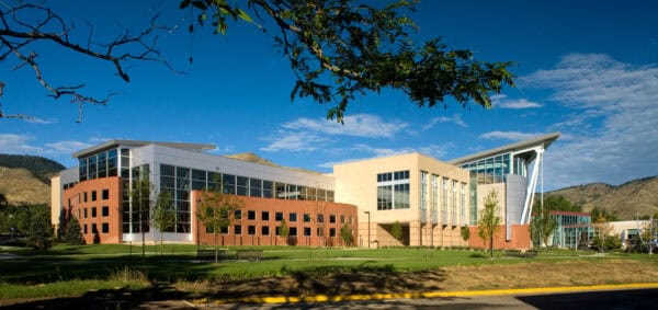 Modern brick-and-glass academic building basks in sunlight, its large windows reflecting blue sky; set on a grassy lawn with benches, framed by tree branches and low hills in the background.