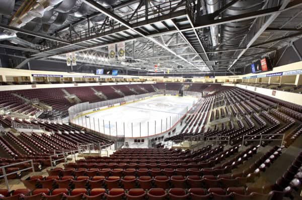 Ice hockey rink sits empty, encircled by protective glass and advertising boards, with rows of maroon seats ascending beneath exposed steel trusses, ducts, banners and bright arena lights.

Transcribed text: "212", "216", "206" (section numbers). Other signage and ribbon-board text in the image are present but not legible at this resolution.