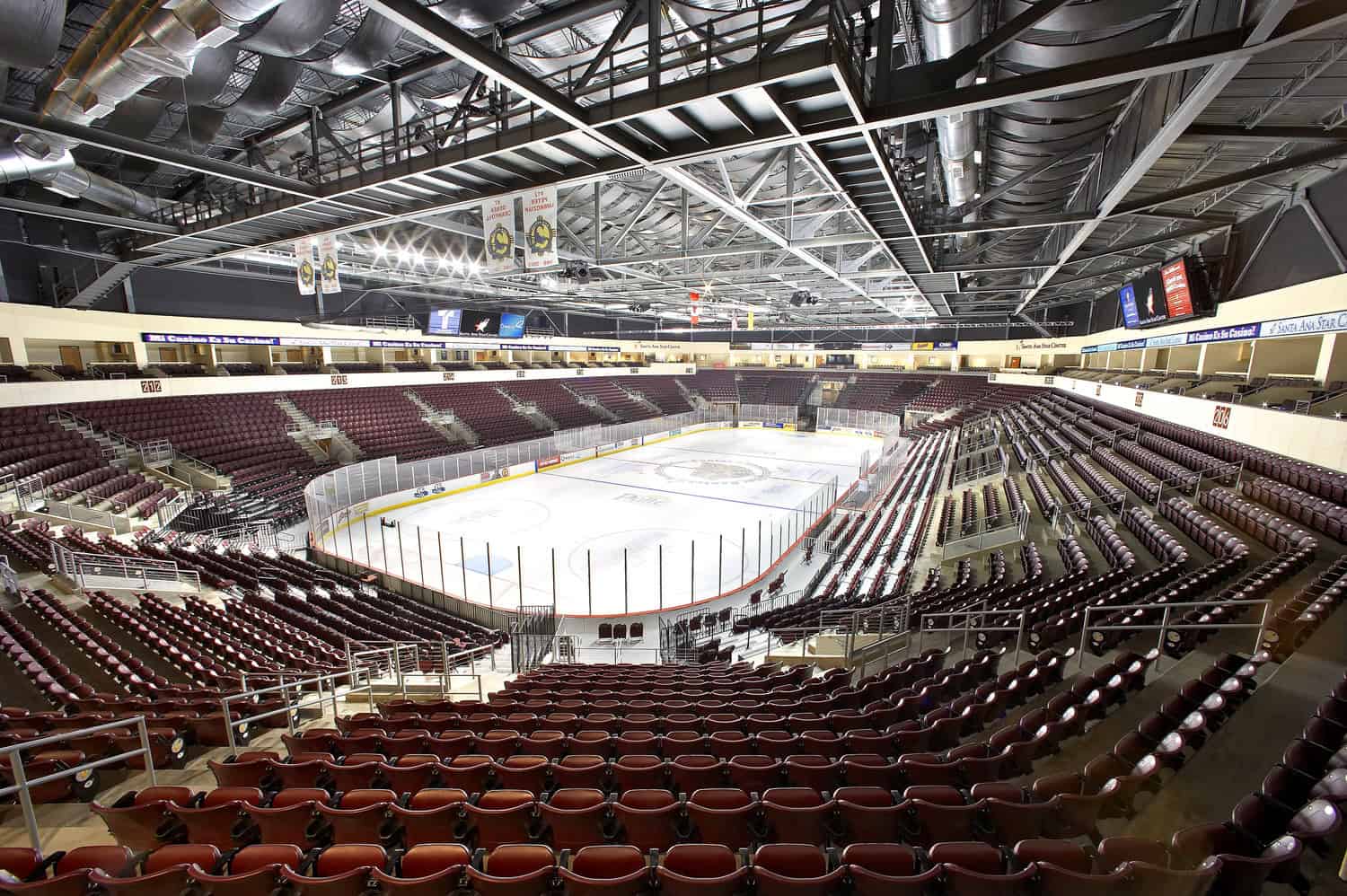 Ice hockey rink sits empty, encircled by protective glass and advertising boards, with rows of maroon seats ascending beneath exposed steel trusses, ducts, banners and bright arena lights.

Transcribed text: "212", "216", "206" (section numbers). Other signage and ribbon-board text in the image are present but not legible at this resolution.