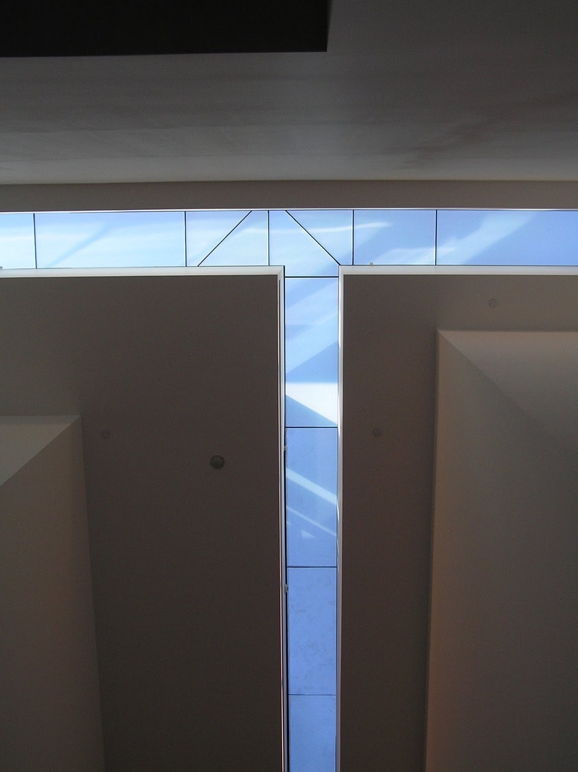 T-shaped skylight (object) admits blue-tinted light and diagonal shadows (action) between two tall recessed ceiling walls in a minimalist interior space (context).