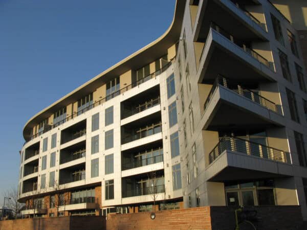 Curved modern apartment building rises with staggered balconies and large glass windows, sunlit on its facade, set against a clear blue sky with a low brick wall and trees foreground.
