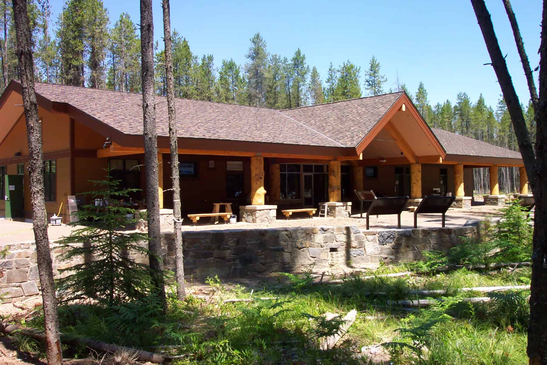 A single-story wooden lodge sits behind a low stone wall, its covered porch supported by log columns with benches and information panels, surrounded by pine trees beneath a clear blue sky.