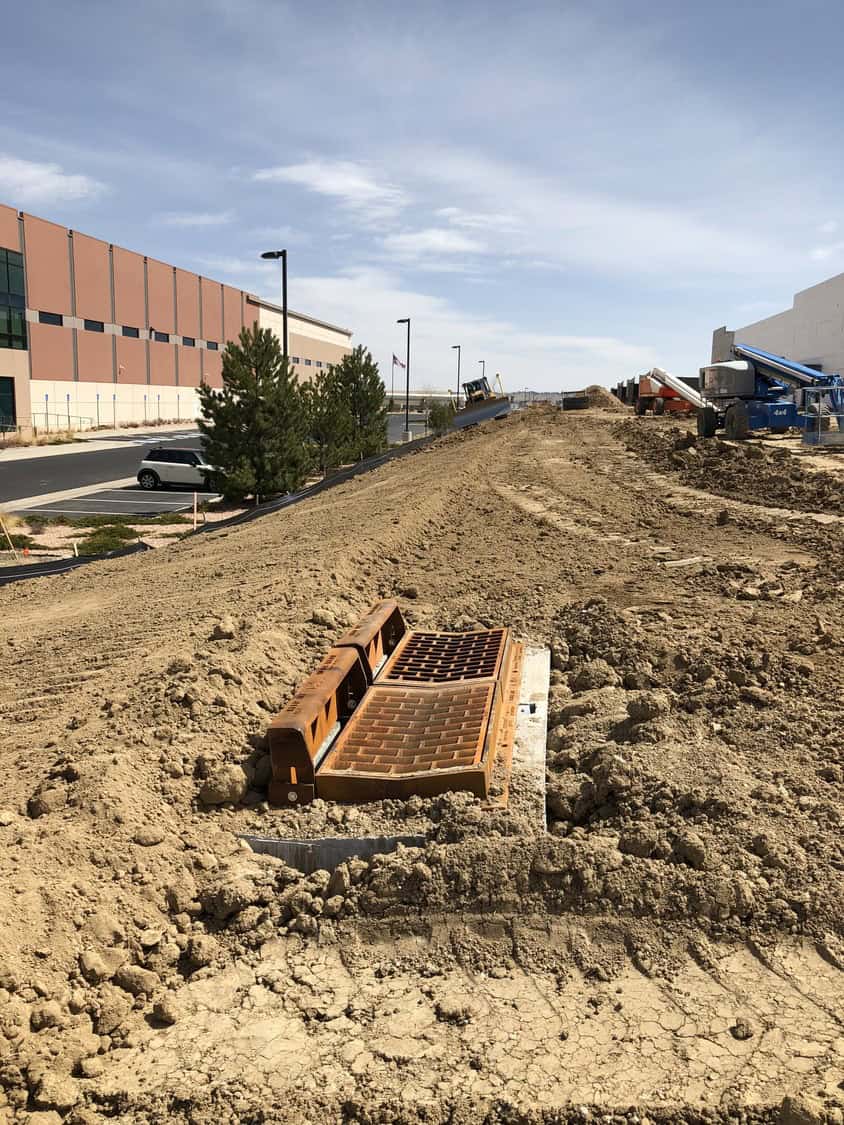 Metal storm drain grate rests partially installed amid freshly graded dirt, surrounded by earth-moving equipment and mounded soil on a construction site beside warehouse buildings, parking lot, and clear sky.