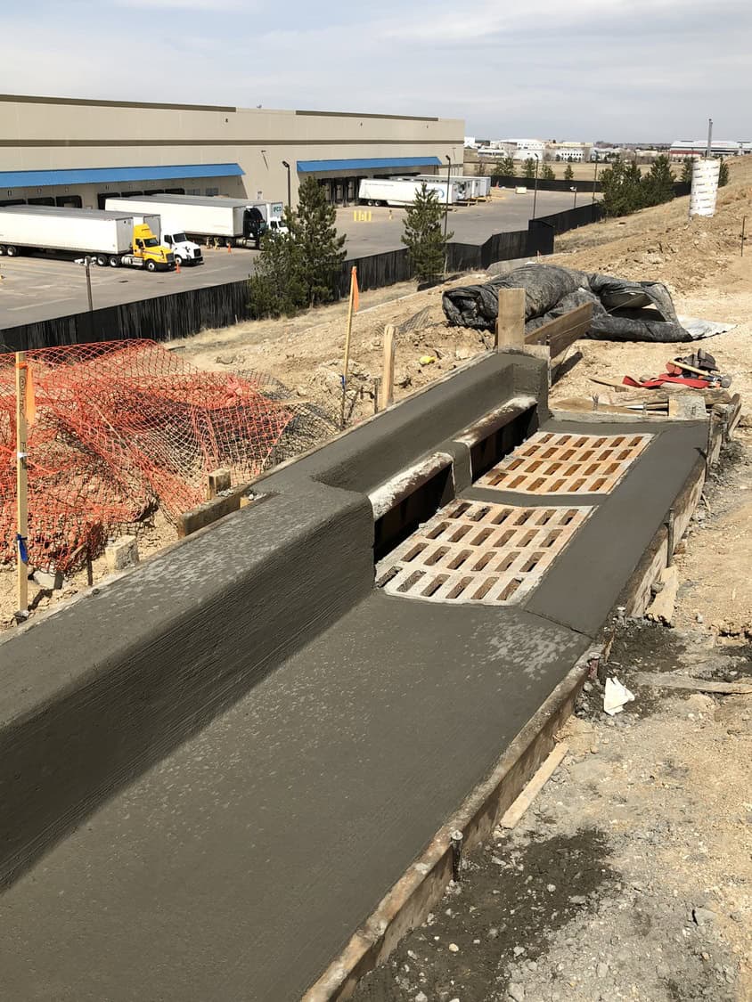 Concrete drainage channel with rectangular metal grates — freshly poured and smoothed — on a dirt construction slope above a warehouse loading-dock area with parked semi-trailers.