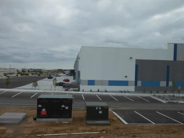 Large white-and-gray warehouse sits beside nearly empty paved parking and loading area; two green electrical transformer boxes occupy the foreground under cloudy sky; sparse young trees and few parked cars.

Text: small labels and warning/warning signs on the transformer boxes are present but illegible at this resolution.