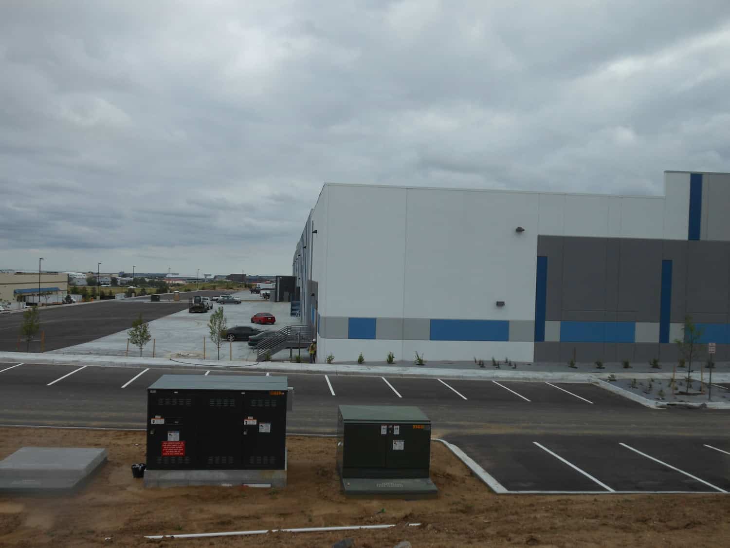 Large white-and-gray warehouse sits beside nearly empty paved parking and loading area; two green electrical transformer boxes occupy the foreground under cloudy sky; sparse young trees and few parked cars.

Text: small labels and warning/warning signs on the transformer boxes are present but illegible at this resolution.