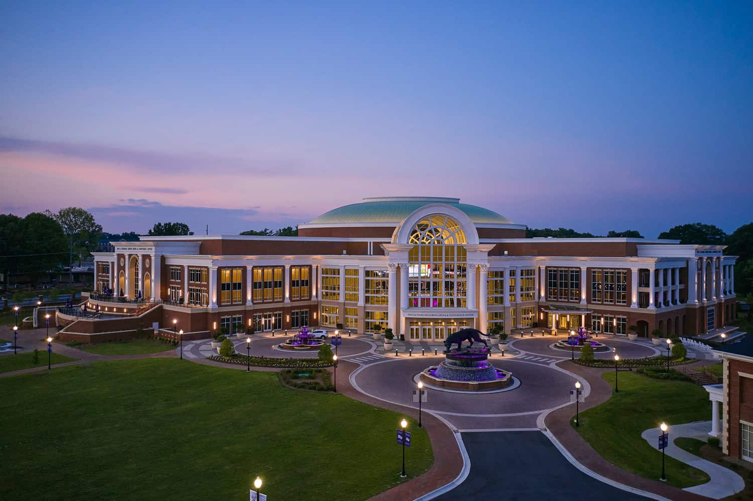 Nido & Mariana Qubein Arena and Conference Center glows with interior lighting and purple-lit fountains in front, framed by curved driveways and manicured lawns at dusk beneath a pastel sky.

Text found: "NIDO & MARIANA QUBEIN ARENA & CONFERENCE CENTER"; banners read "HIGH POINT UNIVERSITY."