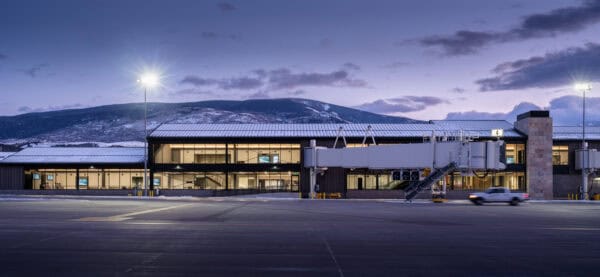 Airport terminal with jet bridge labeled "4" stands idle; a pickup drives past on a snow-dusted apron at dusk, illuminated windows and floodlights, snowy mountains behind.
