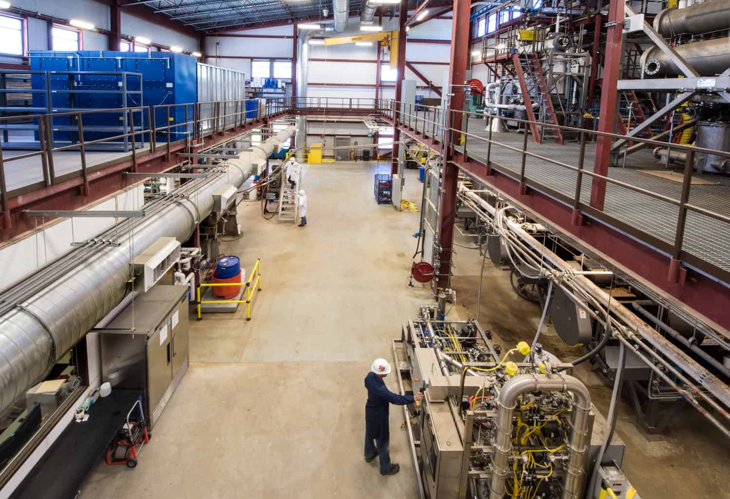 A worker in a hard hat operates a control panel on industrial machinery, while other technicians and large ducts, pipes, tanks and elevated metal walkways occupy a spacious factory hall.