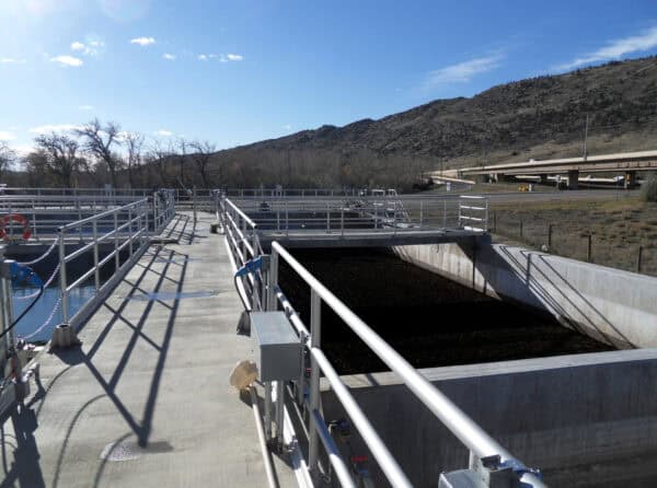 Concrete walkway with metal railings crosses a treatment basin of dark sludge; long shadows fall on the platform, hills and bare trees beside a highway overpass under clear blue sky.
