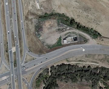 Small rectangular building sits in a dirt-cleared roadside lot beside a curved highway ramp; scattered vehicles and debris dot the site, surrounded by dry grassland and a treed embankment.