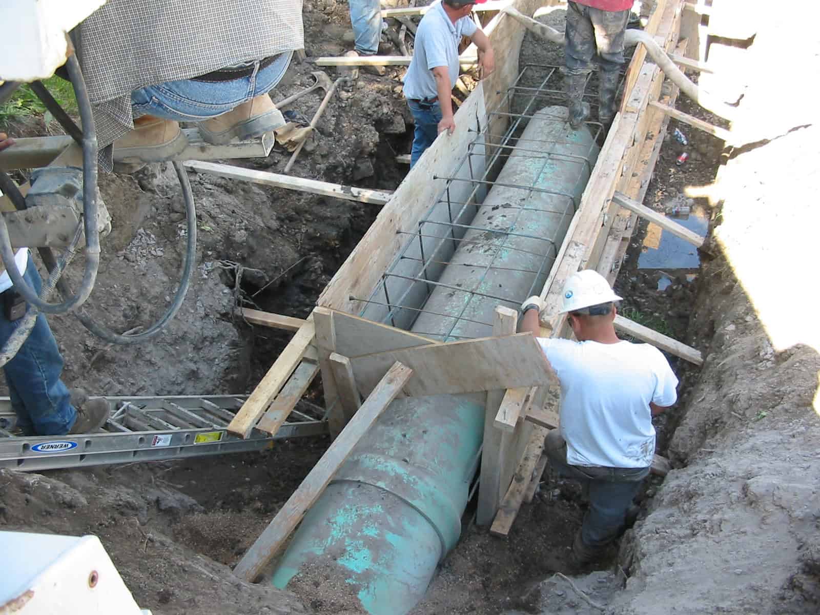 Large green utility pipe; workers secure wooden forms and steel rebar around it inside a deep trench at a muddy construction site. Text: "WERNER" (on ladder).