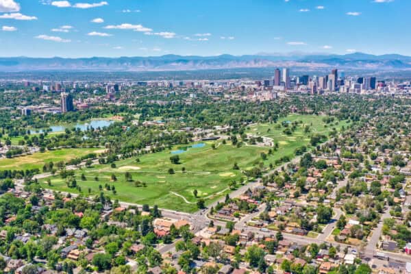 Golf course spreads across green fairways, sand traps and ponds, bordering tree-lined residential streets; downtown skyscrapers rise beyond, with distant mountains under a clear blue sky.