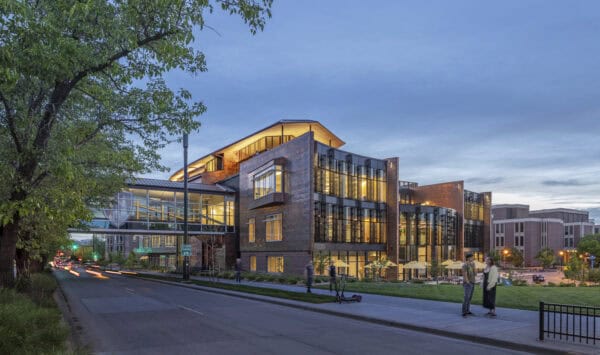 Glass-and-copper academic building glows with interior lights at dusk, connected by a skybridge; pedestrians converse on the sidewalk while cars pass along a tree-lined campus street.