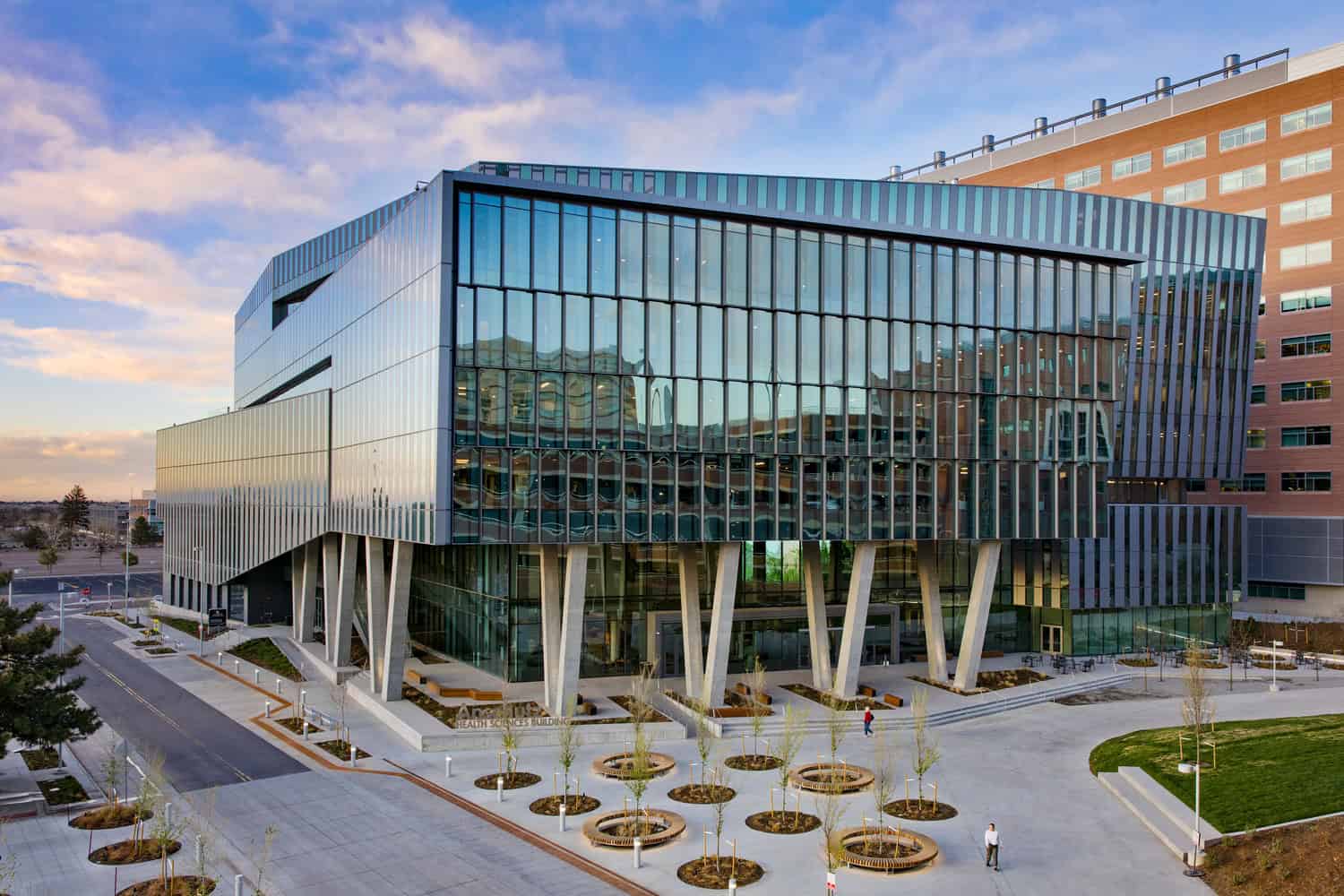 Modern glass-and-steel building rests on angled concrete pillars, reflecting sunset sky and nearby structures, facing a plaza with circular tree planters, scattered pedestrians, and an adjacent brick building.

Text: "Anschutz Health Sciences Building"