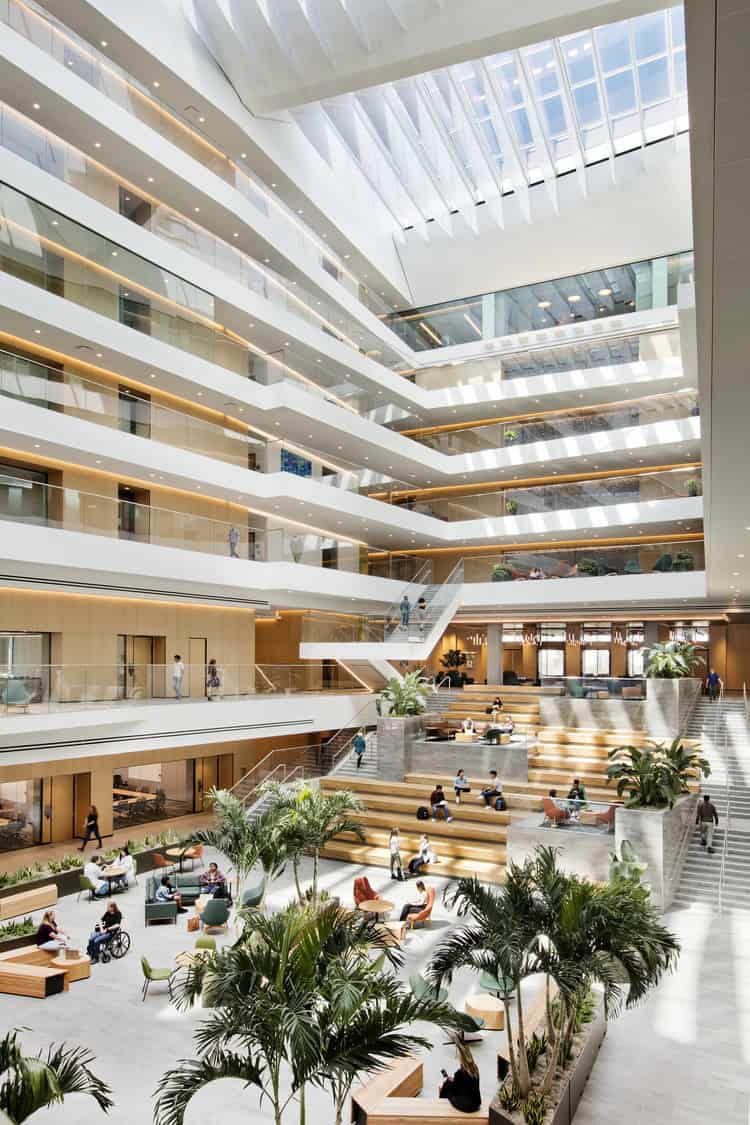 Atrium: tiered wooden seating and tropical plants host people sitting, working on laptops, and walking; surrounded by glass-walled office floors and staircases beneath a large skylight in a modern building.