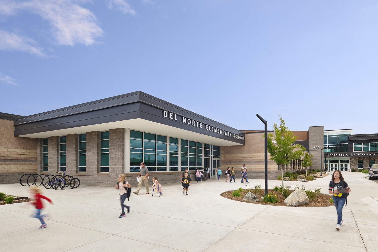Del Norte Elementary School building: children and adults walk and run across a broad concrete plaza toward and away from the glass-front entrance, beside bike racks and planted beds under a clear sky.

Transcribed text: DEL NORTE ELEMENTARY SCHOOL; UPPER RIO GRANDE SCHOOL; 950