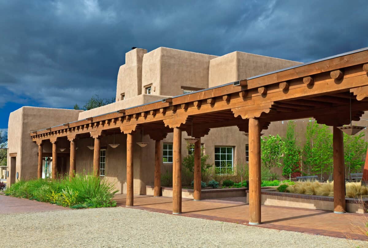 Wooden colonnade extends along an adobe building, supporting a carved-beam porch; sunlight illuminates wooden columns and landscaped desert plants beneath dramatic dark storm clouds.