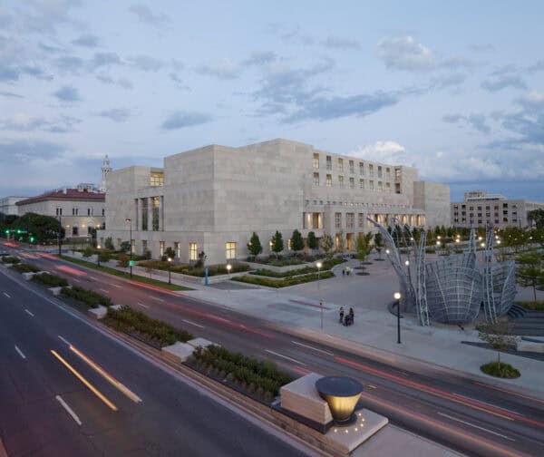 A large rectangular stone civic building sits beside a plaza; cars carve light trails on the adjacent boulevard at dusk, with a metal sculpture and planted trees framing the scene.