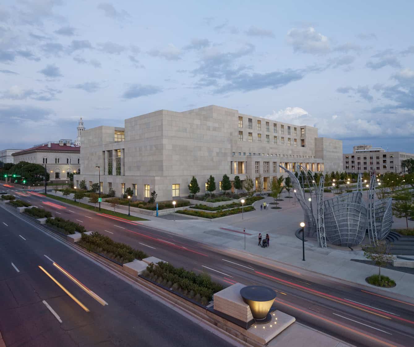 A large rectangular stone civic building sits beside a plaza; cars carve light trails on the adjacent boulevard at dusk, with a metal sculpture and planted trees framing the scene.