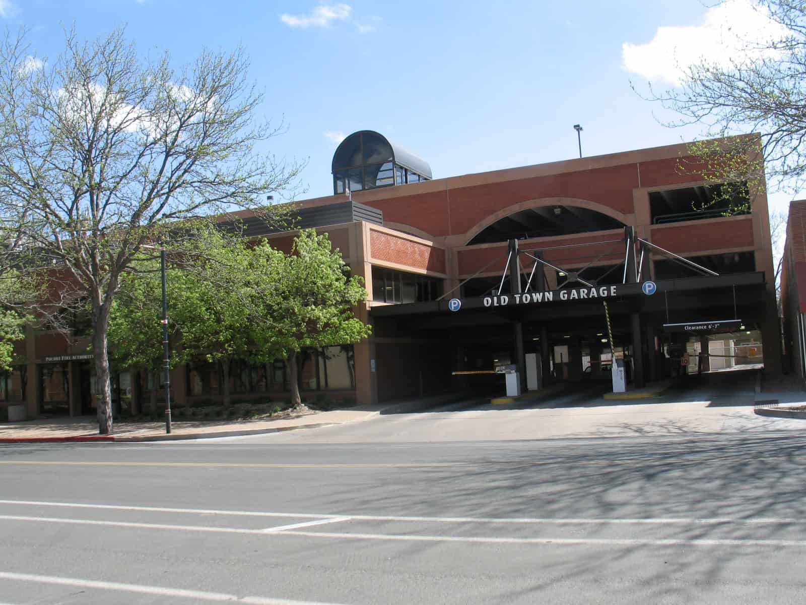 Parking garage entrance labeled "OLD TOWN GARAGE" (P signs visible) sits open with entry lanes and a "Clearance 6'-7"" bar; multi-story brick building flanked by trees under a blue sky.