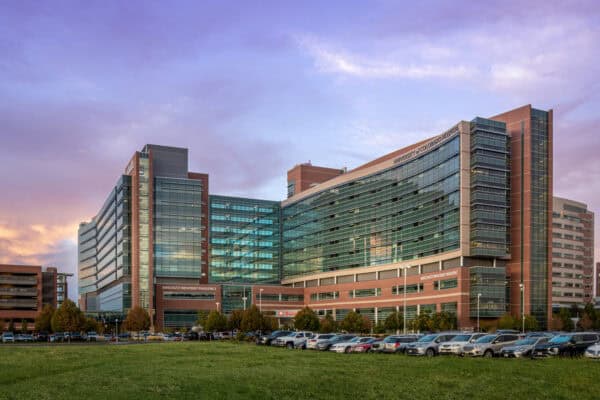 Large glass-and-brick hospital complex stretches across a grassy foreground, its linked glass wings reflecting a purple sunset, with a row of parked cars along the entrance.

Text in image: "UNIVERSITY of COLORADO HOSPITAL" ; "ANSCHUTZ INPATIENT PAVILION 3" ; "ANSCHUTZ INPATIENT PAVILION 2" ; "Emergency"